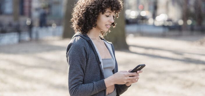 Curly haired young lady looking at her phone while standing outside a sunny day