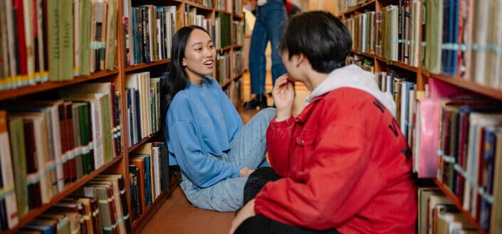 Two youngsters talking to each other while sitting on the library floor