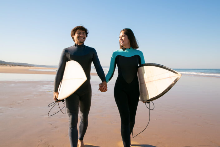 Surfer couple holding hands while walking on the shore.