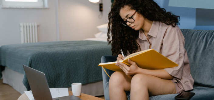 Curly haired woman sitting on a sofa while writing on her yellow journal notebook