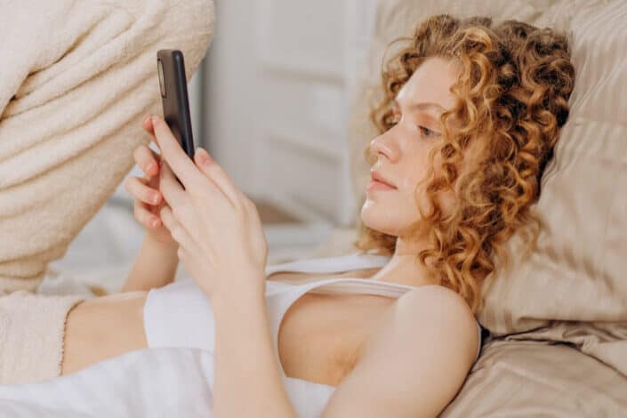 Woman in white tank top lying on bed holding smartphone