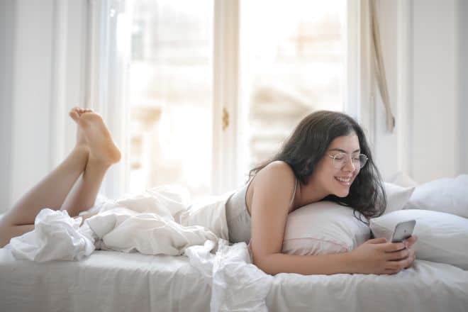 Woman with eyeglasses laying in bed using phone - Questions to Ask Your Crush While Texting